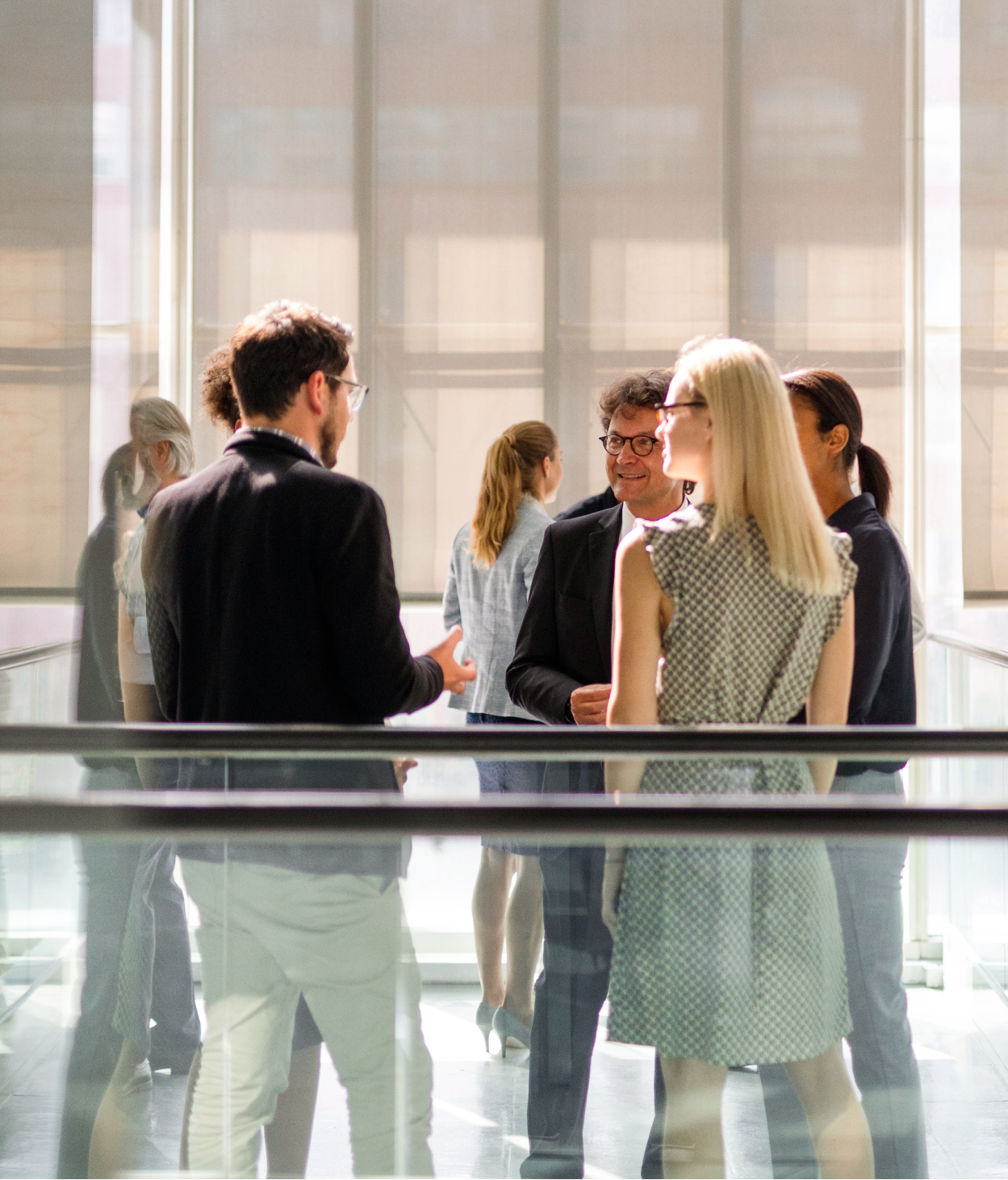 photo, employees chat at a gathering in a large office hallway. They are standing near waist-high glass guard rails with brown banisters. A large building can be seen through the open window nearby.